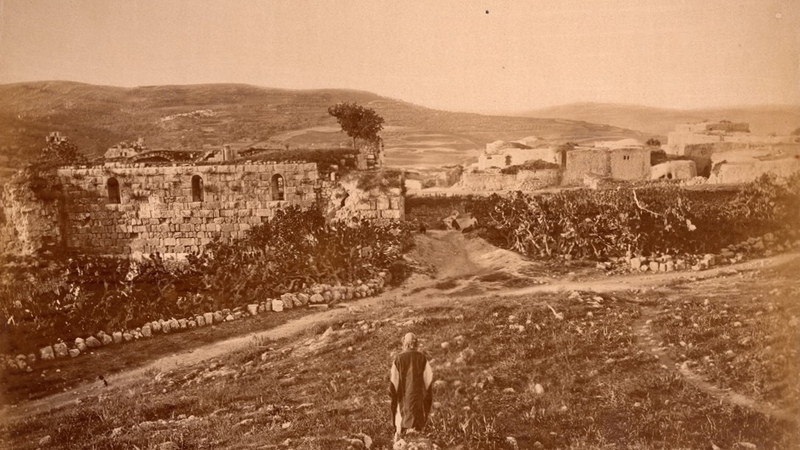 Sepia-toned photograph of ancient stone ruins and surrounding landscape in Samaria (Sebaste), with a person sitting in the foreground. Hills and buildings are visible in the background.