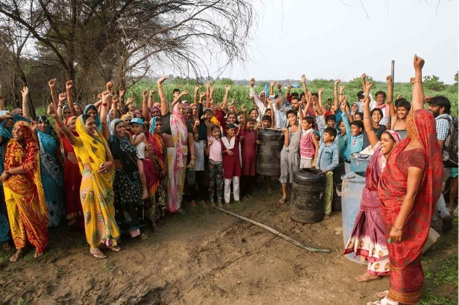 A group of women and children, most them smiling, standing outdoors, raising their hands. They are dressed in colorful traditional attire and gathered beside barrels in a rural setting.