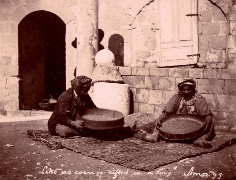 Two people sifting grain outside, in the shade of a building.