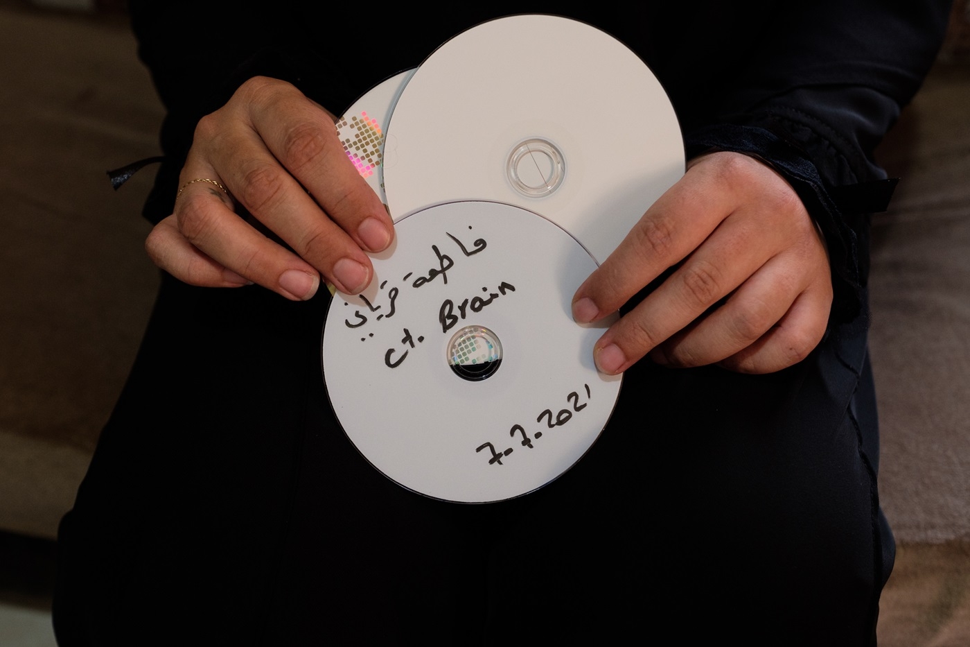 A close-up of hands holding a number of CDs up to the camera; the CD at the front reads: "Fatima Qiryani C.T. Brain 7-7-2021".