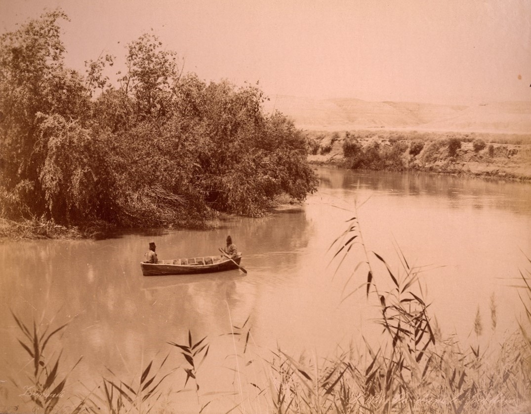 Two people sit in a small rowing boat on a calm river. Trees and bushes line the riverbank, and hills are visible in the background.