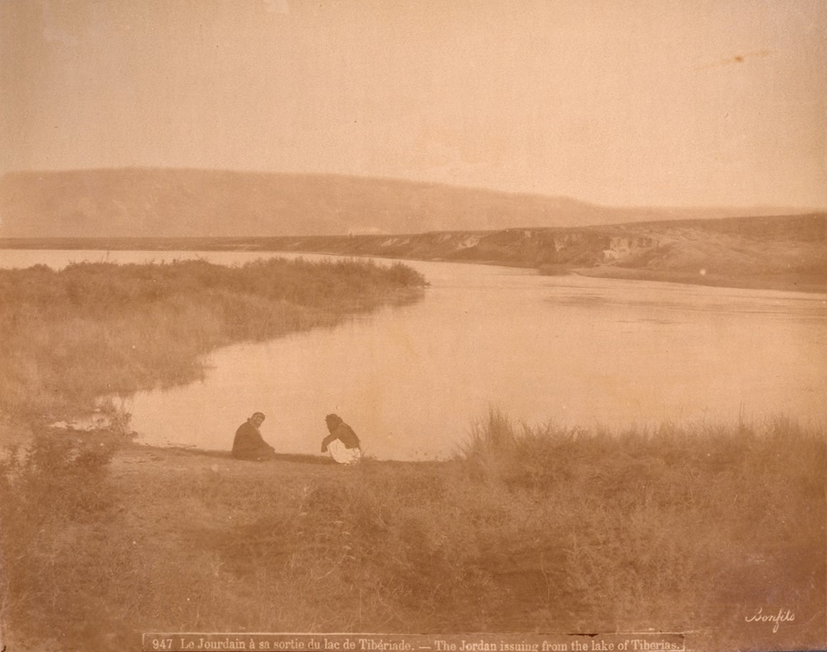 Two people sit at a riverbank. The riverbank is covered in bushes and shrubbery.