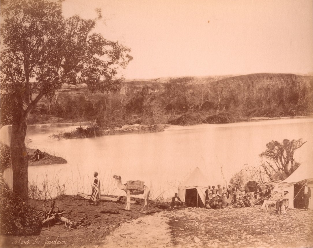 Tents, a group of people, and a camel on a riverbank. Trees and bushes line the riverbank, and hills and mountains are visible in the background.