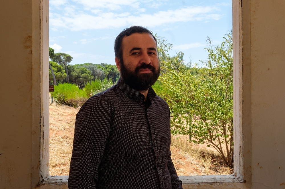 A smiling man stands in front of a window, behind which are trees and shrubbery, and a blue sky.