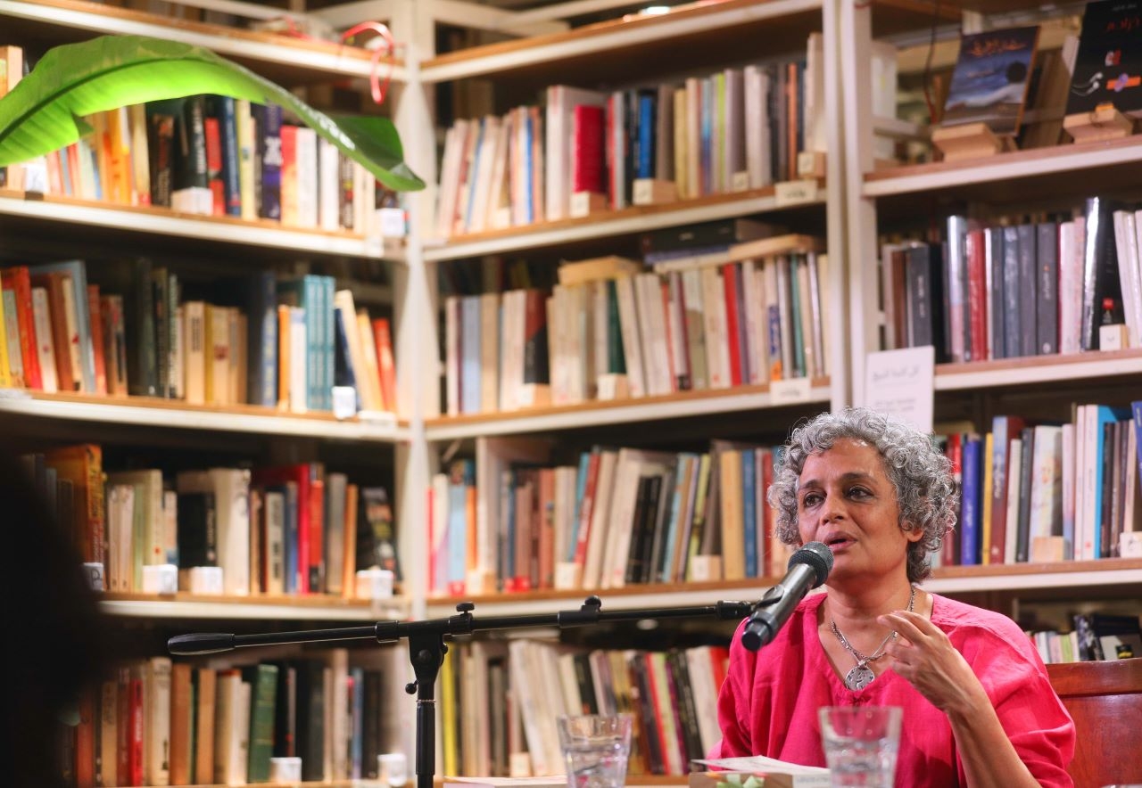 Indian author Arundhati Roy speaks into the microphone at Barzakh Bookshop in Hamra, with a vast array of books visible behind her.