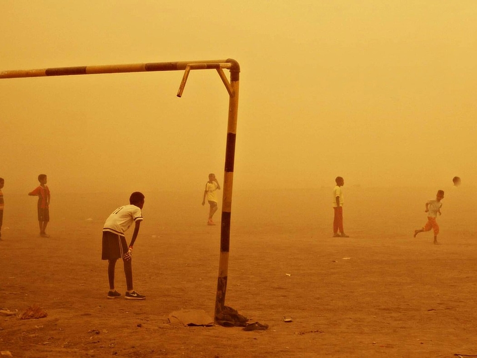 Kids playing football on the sand in the middle of a sandstorm that colors the entire photo golden-yellow.
